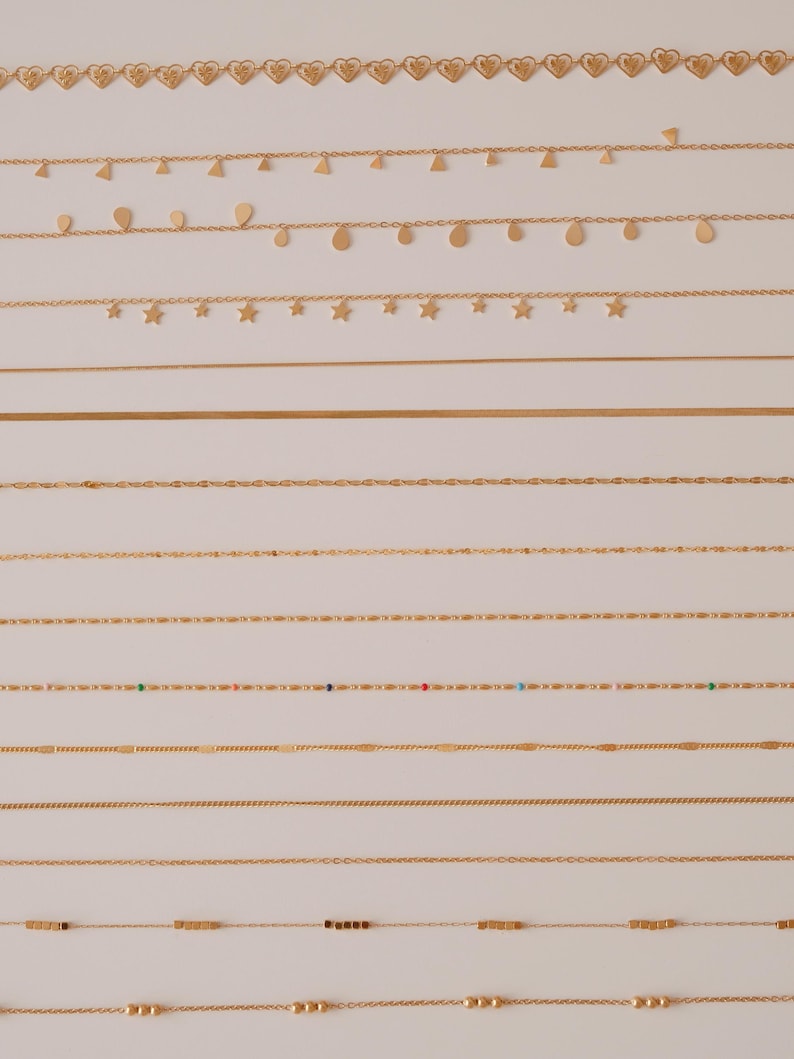K&ouml;nnte beinhalten: Eine Sammlung von 12 goldenen Ketten mit verschiedenen Stilen und Designs. Die Ketten sind in Reihen auf einem wei&szlig;en Hintergrund angeordnet. Einige Ketten haben kleine Anh&auml;nger, wie Herzen, Sterne und Tropfen.