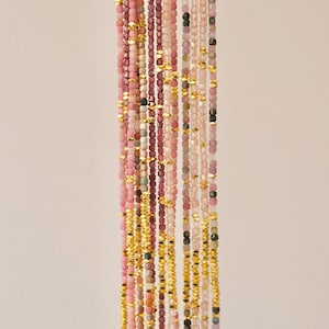 May include: Vertical composition of multiple strands of gemstone beads in shades of pink, purple, and green, interspersed with gold-colored metal beads. The beads are small and square-shaped, creating a delicate, elegant aesthetic.