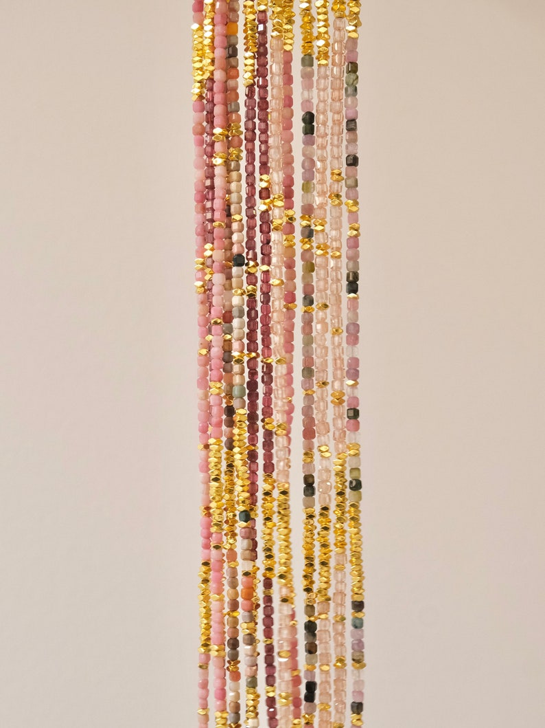 May include: Vertical composition of multiple strands of gemstone beads in shades of pink, purple, and green, interspersed with gold-colored metal beads. The beads are small and square-shaped, creating a delicate, elegant aesthetic.