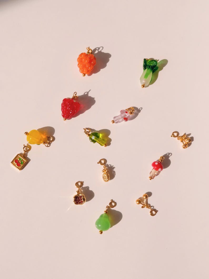 K&ouml;nnte beinhalten: Eine Sammlung von 12 bunten Glasanh&auml;ngern, darunter eine Ananas, eine Birne, ein Pilz, eine Himbeere, eine Kirsche, eine Traube, eine Zitrone, ein gr&uuml;nes Blatt, eine Erdbeere und eine kleine Schachtel mit einer Kirsche darin. Die Anh&auml;nger sind alle vergoldet und haben ein zartes, verspieltes Design.
