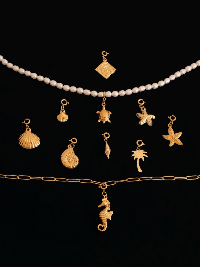 K&ouml;nnte beinhalten: Eine goldene Kettenkette mit einer Vielzahl von goldenen Anh&auml;ngern, darunter ein Seepferdchen, eine Palme, eine Muschel, ein Seestern, eine Schildkr&ouml;te und eine spiralf&ouml;rmige Muschel. Die Anh&auml;nger h&auml;ngen an einer goldenen Kettenkette.
