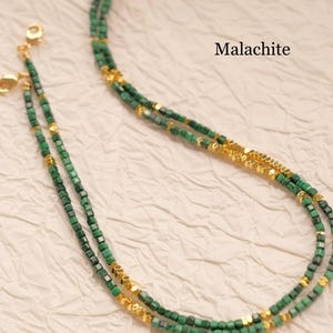 May include: A layered necklace featuring small, square, green malachite beads interspersed with gold-colored accents. The necklace is displayed on a textured, off-white surface. The word "Malachite" is printed in black.