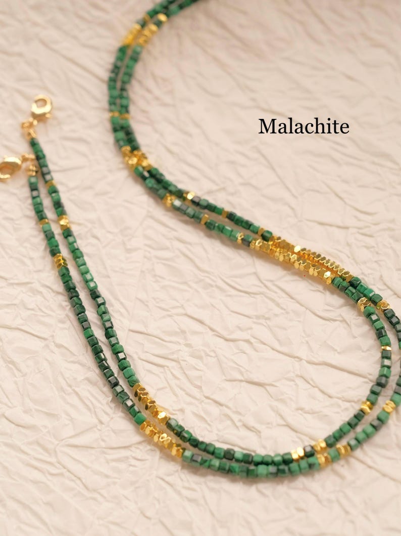 May include: A layered necklace featuring small, square, green malachite beads interspersed with gold-colored accents. The necklace is displayed on a textured, off-white surface. The word "Malachite" is printed in black.