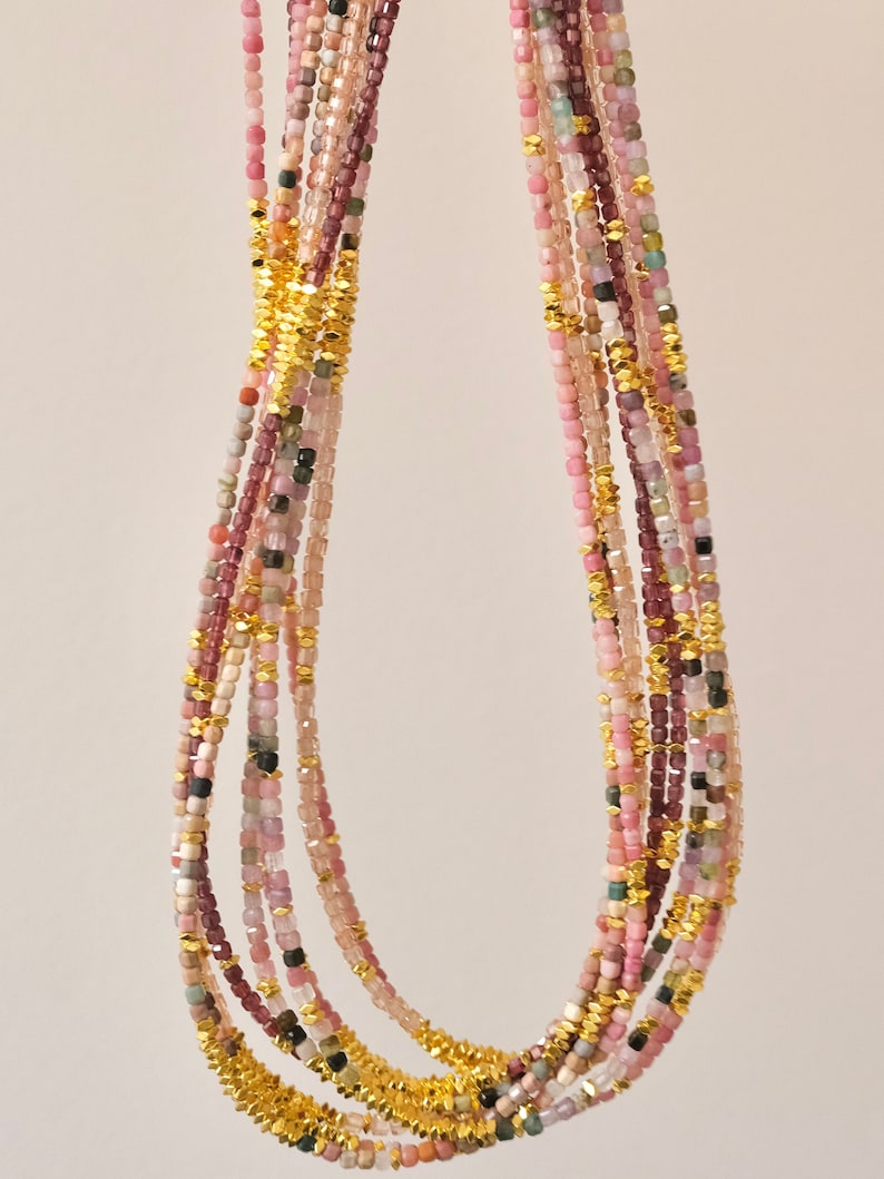 May include: A multi-strand necklace featuring small, square-cut gemstones in shades of pink, purple, and green. The strands are interspersed with gold-colored, textured metal accents. The necklace is displayed against a plain, light background.