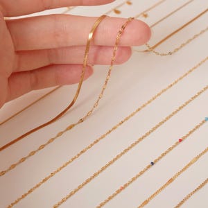 K&ouml;nnte beinhalten: Eine Sammlung von goldenen Kettenhalsketten mit verschiedenen Gliederarten, darunter eine flache Schlangenkette, eine zarte ovale Kette und eine Kette mit kleinen bunten Perlen.