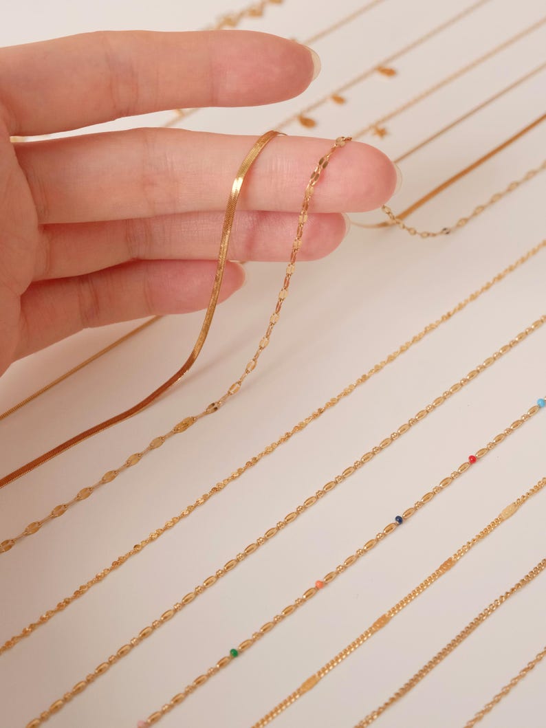 K&ouml;nnte beinhalten: Eine Sammlung von goldenen Kettenhalsketten mit verschiedenen Gliederarten, darunter eine flache Schlangenkette, eine zarte ovale Kette und eine Kette mit kleinen bunten Perlen.