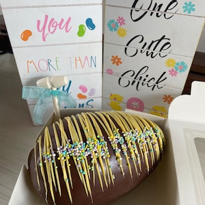 Puede incluir: Un huevo de Pascua de chocolate decorado con glaseado amarillo y chispas de colores. El huevo está en una caja blanca con un letrero que dice "Te amo más que" y otro letrero que dice "Un pollito lindo".