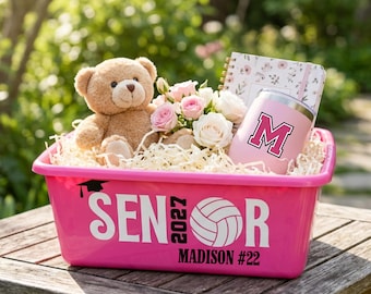 May include: A bright pink plastic bin labeled SENIOR 2027 with a volleyball graphic and the name MADISON #22. Inside are a tan teddy bear, a small bouquet of pink and white roses, a floral patterned notebook, and a pink tumbler with a letter M.