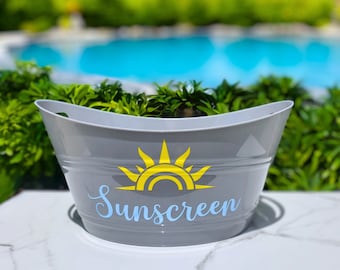 May include: A gray oval metal bucket with a yellow sun graphic and the word Sunscreen written in light blue script lettering. The bucket features horizontal ribbed detailing along the sides and integrated handles on the left and right rims.