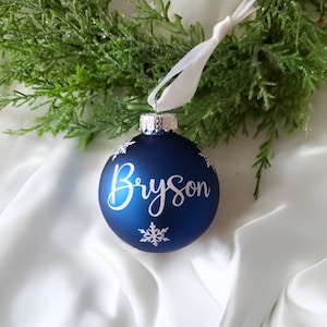 May include: Blue Christmas ornament with white snowflakes and the name "Bryson" written in white cursive.