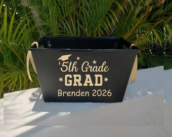 May include: A black metal graduation basket with gold accents and handles. The front of the basket features the text "5th Grade GRAD Brenden 2026" in a gold color, along with a graduation cap and stars.