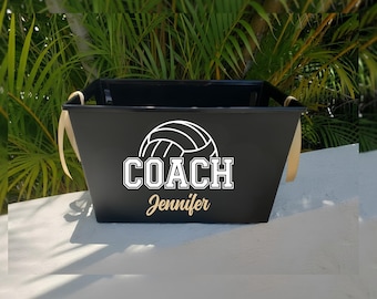 May include: A black rectangular storage bin featuring a white graphic of a volleyball above the word COACH in block lettering and the name Jennifer in a gold script font. The bin has light-colored ribbon handles attached to the sides.