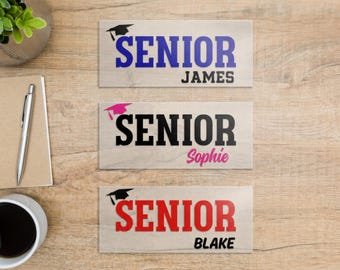 May include: Three rectangular signs with a light wood grain finish. Each sign features a graduation cap icon and the word SENIOR. The first sign has blue text and the name JAMES. The second has black text and the name Sophie. The third has red text and the name BLAKE.
