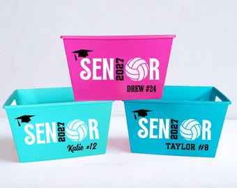以下が含まれることがあります： 鮮やかなピンクとターコイズの長方形のプラスチック製収納ボックス3個。各ボックスには、黒い卒業帽のアイコン、SENIOR 2027の文字、白いバレーボールのグラフィック、そして背番号付きのパーソナライズされた名前（DREW #24、KATIE #12、TAYLOR #8）がデザインされています。