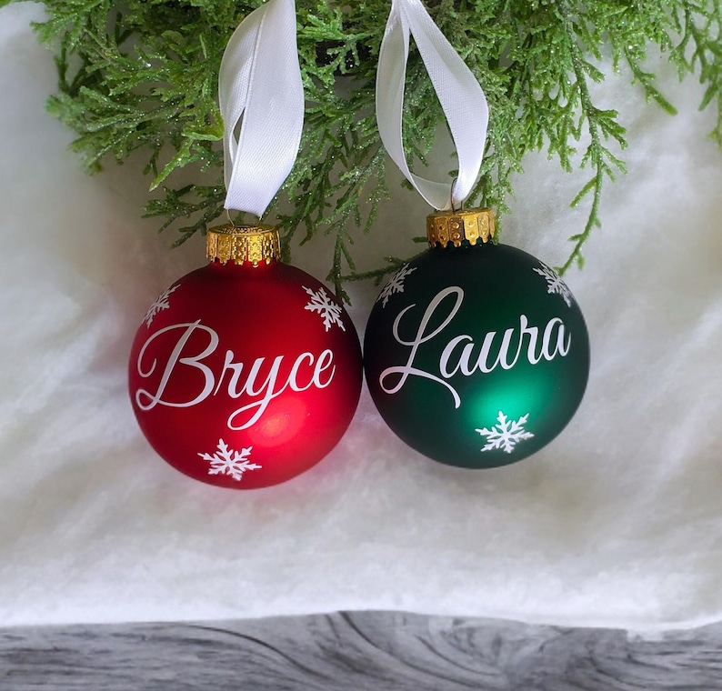 May include: Two personalized Christmas ornaments, one red with the name "Bryce" and one green with the name "Laura", both with white snowflake designs.