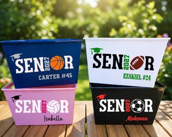 May include: Four rectangular plastic storage bins in blue, white, pink, and black. Each bin features the text SENIOR 2027, a graduation cap icon, a sports ball graphic, and a personalized name with a jersey number printed on the front surface.