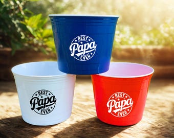 May include: Three plastic cups in blue, white, and red, stacked on a wooden surface. Each cup has the words "Best Papa Ever" in a white circular design. The cups are empty and ready to be filled.