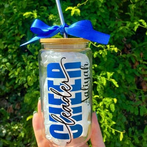 Puede incluir: Un vaso de vidrio transparente con tapa de bamb&uacute; y pajita de metal plateado. Una cinta de sat&eacute;n azul brillante est&aacute; atada alrededor de la pajita. El vaso presenta la palabra CHEER en letras azules negritas y el nombre Aaliyah en cursiva negra.