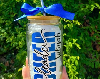 May include: A clear glass tumbler with a bamboo lid and a silver metal straw. A bright blue satin ribbon is tied around the straw. The glass features the word CHEER in bold blue letters and the name Aaliyah in black cursive script text.