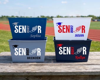 May include: Four rectangular storage bins in navy blue, white, light blue, and dark blue. Each bin features the text SENIOR 2027 with a graduation cap icon and a winged shoe graphic. The names Sophia, Jackson, Brenden, and Kaillyn are printed below.