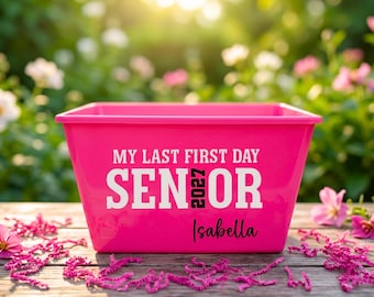 May include: A bright pink plastic bin featuring white text that reads MY LAST FIRST DAY and SENIOR, with the number 2027 in black and the name Isabella in black script. The bin has a wide, open top and a rectangular shape with tapered sides.