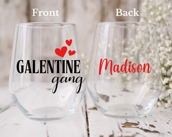 以下が含まれることがあります： 2つのステムレスワイングラス。片方のグラスには「GALENTINE gang」の文字と3つの赤いハートが描かれています。もう一方のグラスには「Madison」という名前が赤色で書かれています。グラスは透明で、白い木の表面に置かれています。