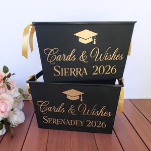 May include: Two black metal graduation card holders with gold text and accents. Each container has a gold graduation cap graphic and the words "Cards & Wishes" and a name and year. Gold ribbons are attached to the sides.