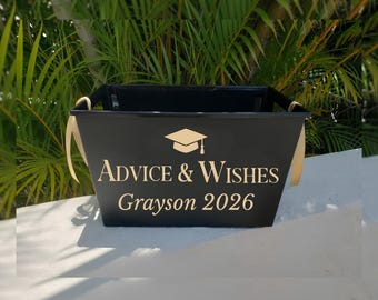 May include: A black rectangular bin featuring gold lettering that reads Advice & Wishes, Grayson 2026, and a gold graduation cap icon. The container has two side handles with light-colored ribbons attached to each side for decorative purposes.