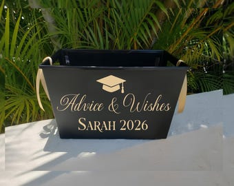 May include: A black metal graduation advice and wishes bucket with gold text and ribbon handles. The text includes a graduation cap graphic, the words "Advice & Wishes", and the name "SARAH 2026".