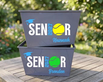 May include: Two gray plastic storage bins stacked on each other. Each bin features a light blue graduation cap, the white text SENIOR, the light blue year 2027, and a name in light blue script. The top bin has a yellow tennis ball, the bottom a green one.