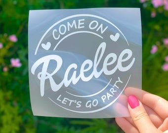 May include: A white vinyl decal on a clear backing sheet featuring the text COME ON Raelee LET'S GO PARTY arranged in a circular design with two small heart icons. The decal is printed in a bold, clean font with a script style for the name.