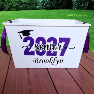May include: A white plastic storage bin with purple ribbon handles. The front features a black graduation cap icon above the purple text 2027, the black script word senior, and the black name Brooklyn printed in a decorative serif font.