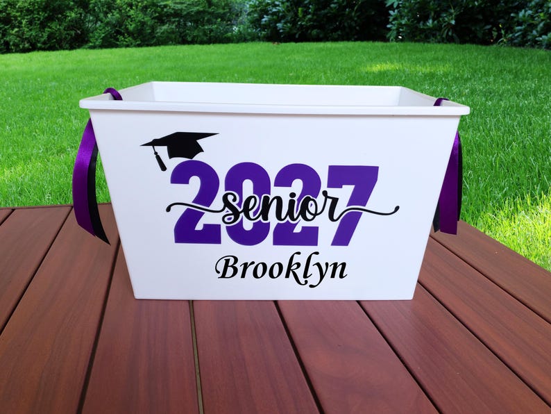 May include: A white plastic storage bin with purple ribbon handles. The front features a black graduation cap icon above the purple text 2027, the black script word senior, and the black name Brooklyn printed in a decorative serif font.