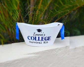 May include: A white oval metal tub featuring blue ribbon handles on each side. The front is printed with a black graduation cap icon above the text "Emma's COLLEGE Survival Kit" in a combination of black and blue lettering.