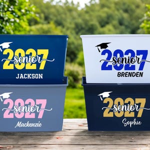 May include: Four rectangular storage bins in navy, white, light blue, and dark blue. Each bin features a graduation cap icon, the year 2027, the word senior in script, and a personalized name: Jackson, Brenden, Mackenzie, or Sophie in various colors.