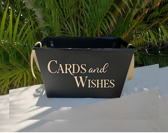 May include: A black rectangular storage bin with gold lettering that reads CARDS and WISHES on the front. The container features two light-colored ribbon handles attached to the sides, providing a simple and functional design for collecting items.