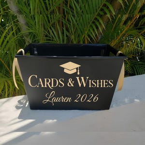 May include: A black metal container with gold ribbon handles. The front of the container has the words "Cards & Wishes" and "Lauren 2026" in gold lettering, along with a graduation cap symbol. The container is set against a backdrop of green foliage.