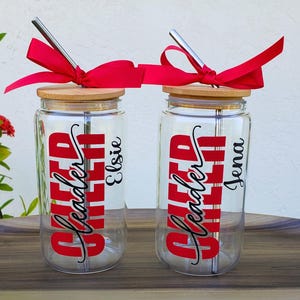 Puede incluir: Dos vasos de cristal transparente con tapas de bamb&uacute; y pajitas de metal. Cada uno tiene un lazo rojo, el texto rojo "Cheer Leader" en fuente negrita y un nombre en letra cursiva negra, "Elsie" en uno y "Jena" en el otro, impresos en el cristal.