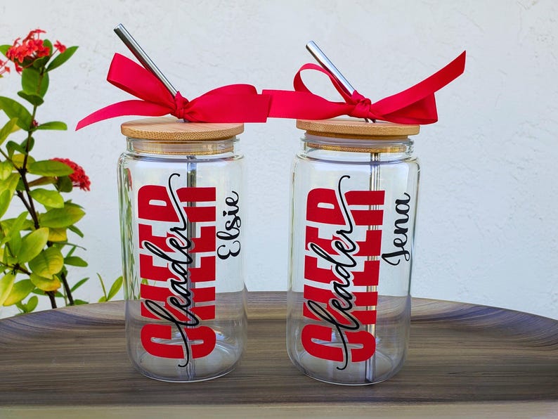 Puede incluir: Dos vasos de cristal transparente con tapas de bamb&uacute; y pajitas de metal. Cada uno tiene un lazo rojo, el texto rojo "Cheer Leader" en fuente negrita y un nombre en letra cursiva negra, "Elsie" en uno y "Jena" en el otro, impresos en el cristal.
