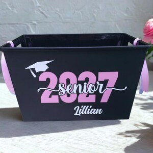 May include: A black rectangular plastic storage basket with light pink ribbon handles. The front features a white graduation cap icon, the pink text 2027, the white cursive word senior, and the white name Lillian printed on the center surface.