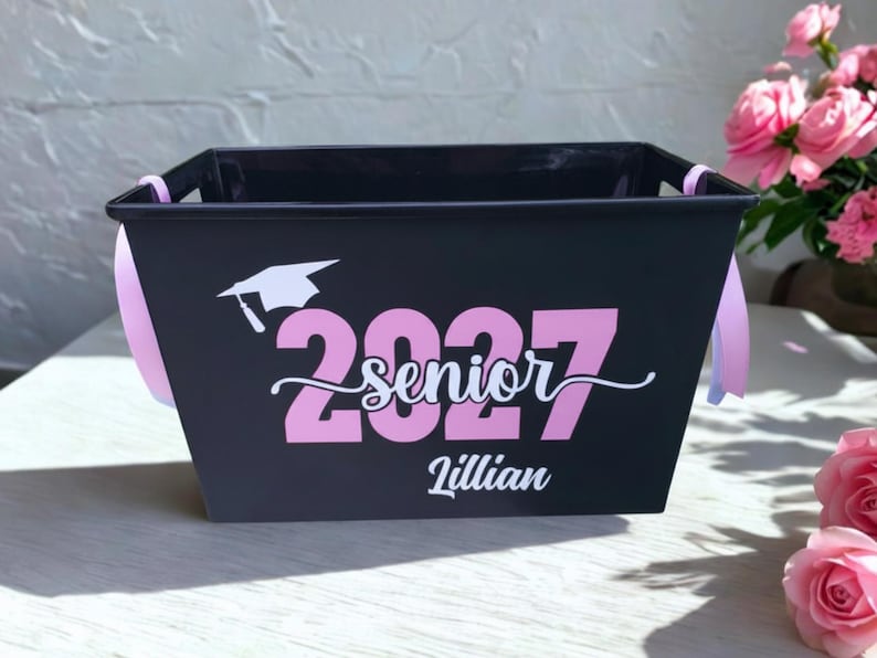 May include: A black rectangular plastic storage basket with light pink ribbon handles. The front features a white graduation cap icon, the pink text 2027, the white cursive word senior, and the white name Lillian printed on the center surface.