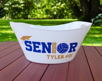 May include: White metal tub with a curved design, featuring the word "SENIOR" in blue, the year "2026" and a volleyball graphic. The name "Tyler #21" is printed in gold. A graduation cap graphic is also visible.