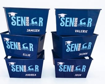 May include: Six navy blue rectangular bins stacked on top of each other. Each bin has a white graduation cap, the word "SENIOR 2026", and a swimmer graphic. Each bin has a different name in white: Jamisen, Valerie, Ellie, Jasmin, Andrea, and Josh.