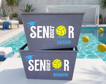 May include: Two stacked gray plastic storage bins featuring white text that reads SENIOR with the year 2027 vertically between the letters. A yellow volleyball replaces the letter O, and a light blue graduation cap sits above the word.