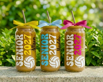 May include: Three glass can-shaped tumblers filled with iced coffee, each featuring a volleyball graphic, the text "SENIOR 2027," and a personalized name. Each tumbler is topped with a metal straw and a matching yellow, blue, or pink ribbon bow.