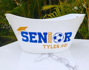 May include: White oval-shaped container with a graduation cap graphic and the word "SENIOR" in blue, "2026" in gold, and a soccer ball graphic. The name "TYLER #21" is also in gold. The container is likely for a graduation or sports-related event.