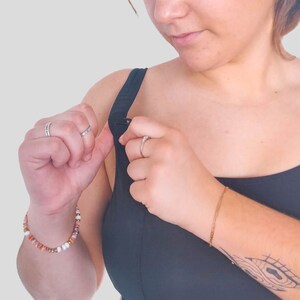 Op de afbeelding: Een vrouw past de sluiting van een zwarte sportbeha met racerback-design aan. Ze draagt een kralenarmband om haar linker pols en een gouden armband om haar rechter pols.