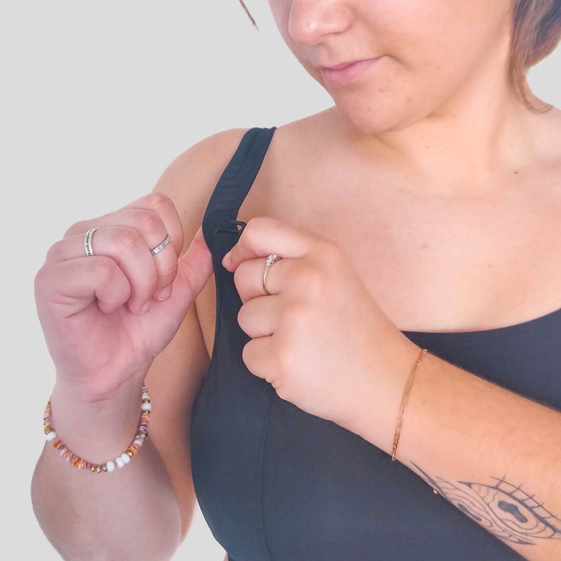 Op de afbeelding: Een vrouw past de sluiting van een zwarte sportbeha met racerback-design aan. Ze draagt een kralenarmband om haar linker pols en een gouden armband om haar rechter pols.