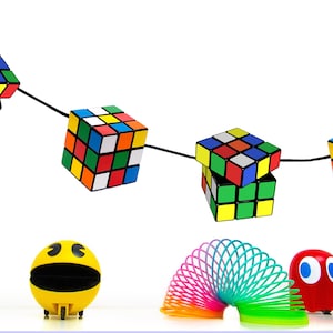 May include: A string of colourful Rubik's Cubes hangs from a black cord. Below, there are a yellow Pac-Man toy, a rainbow slinky, a red ghost toy, and two smaller Rubik's Cubes.