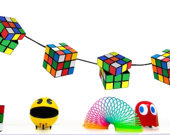 May include: A string of colorful Rubik's Cubes hangs from a black cord. Below, there are a yellow Pac-Man toy, a rainbow slinky, a red ghost toy, and two smaller Rubik's Cubes.